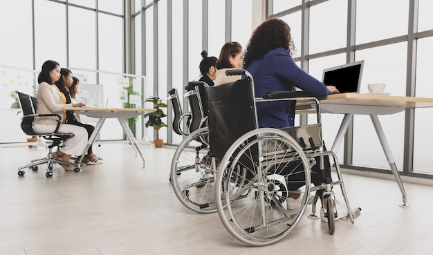 Asian business women including handicap woman sitting on wheelchair working hard on laptop on the table in office. Concept for business meeting.