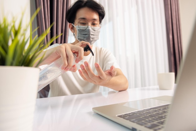 Asian business young handsome man glasses working from home office he quarantines disease coronavirus or COVID-19 wearing protective mask and cleaning hands with sanitizer gel on front laptop computer