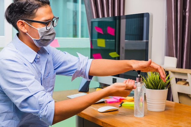 ホームオフィスで働くアジアのビジネスの若い男性は、病気のコロナウイルスまたはCOVID-19を隔離し、保護マスクを着用し、フロントPCコンピュータで消毒ジェルで手を掃除します