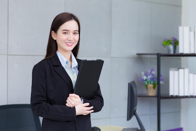 Asian business young woman in black suit smiles happily while she works and holds clipbroad