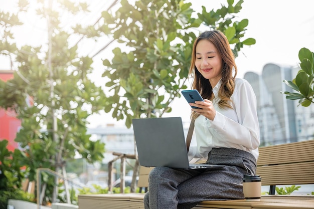 Asian business young woman working laptop and using mobile smartphone outdoor building exterior, Happy smiling businesswoman sit writing text on mobile phone outside street city near office in morning