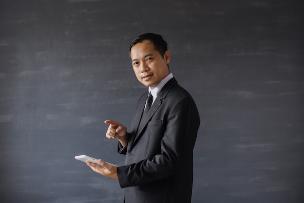 Uomo d'affari asiatico in abito nero che guarda la telecamera mentre indica lo smartphone
