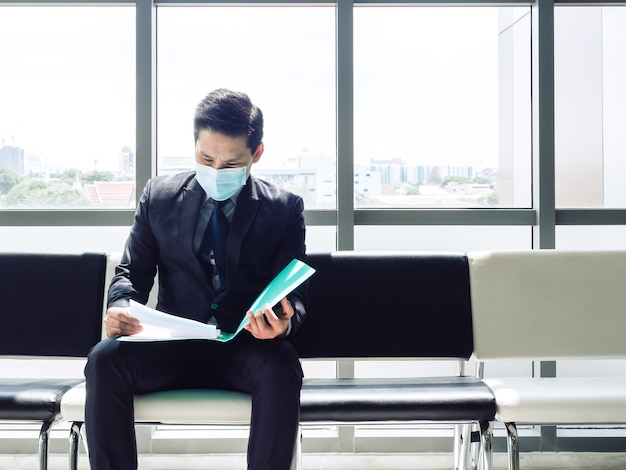 黒いスーツを着たアジア人ビジネスマンが保護マスクを着用して座って履歴書を確認し、巨大なガラス窓の近くで就職の面接を待っています