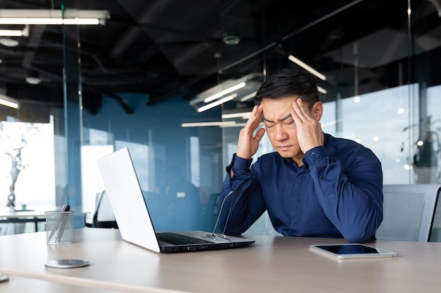 Asian businessman boss sick has severe headache man in glasses sitting at desk using laptop at work