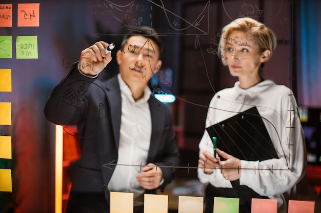 Asian businessman brainstorming talking with female colleague standing behind glass board