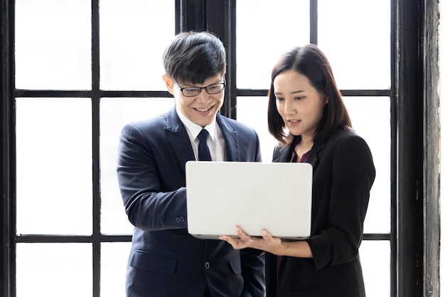 Азиатский бизнесмен и деловая женщина запускают вместе компьютерный ноутбук, стоя в окнах офисного здания Успешные молодые деловые партнеры в костюмах смотрят на стратегический анализ ноутбука