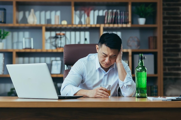 아시아 사업가는 절망에 빠진 남자가 우울하고 희망이 없는 사무실의 테이블에 늦게 앉아 독주를 마신다