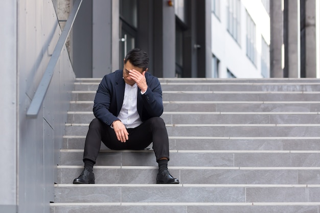 Asian businessman has a severe headache sitting on the stairs near the office