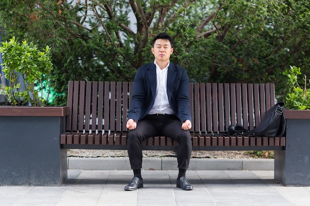 Asian businessman performing breathing exercises trying to calm\
stress, sitting on a bench during a lunch break in a business\
suit