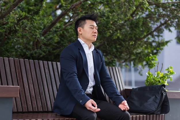 Asian businessman performing breathing exercises trying to calm\
stress, sitting on a bench during a lunch break in a business\
suit