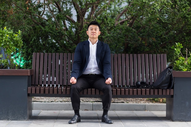 Asian businessman performing breathing exercises trying to calm stress, sitting on a bench during a lunch break in a business suit