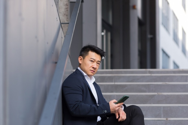 Photo asian businessman reads bad news from the phone, sitting near the office on the stairs, depressed, losing hope