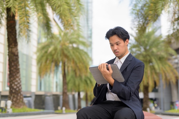 Asian businessman sitting outdoors in city using digital tablet computer