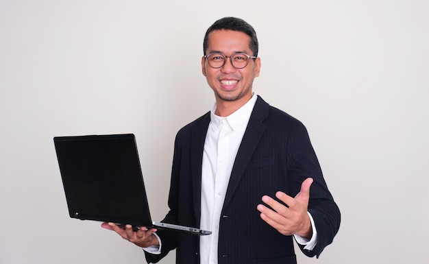 Photo asian businessman smiling and showing inviting hand gesture while holding a laptop
