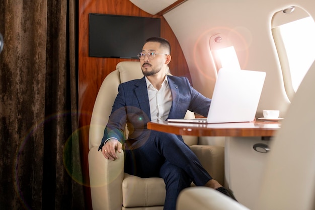 Asian businessman in suit and glasses flies in private luxury plane with laptop Korean trader