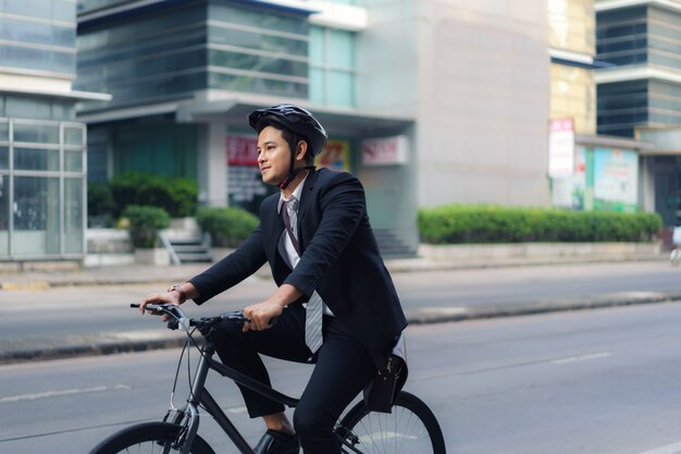 スーツを着たアジア人ビジネスマンが、朝の通勤のために街の通りを自転車で走っています。エコ輸送コンセプト。