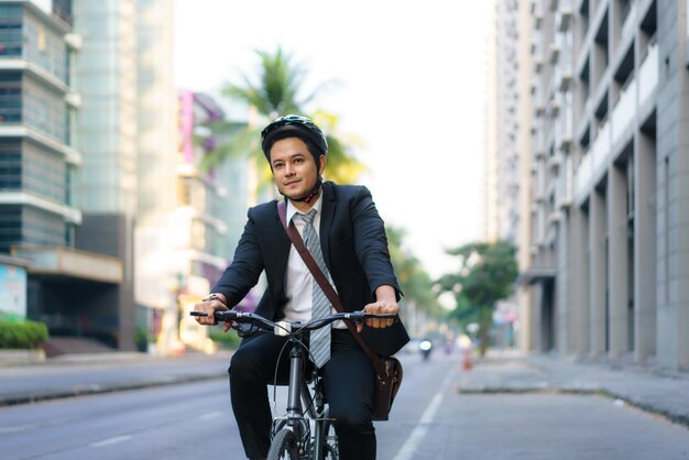 スーツを着たアジア人ビジネスマンが、朝の通勤のために街の通りを自転車で走っています。エコ輸送コンセプト。