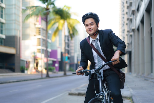 スーツを着たアジア人ビジネスマンが、朝の通勤のために街の通りを自転車で走っています。エコ輸送コンセプト。