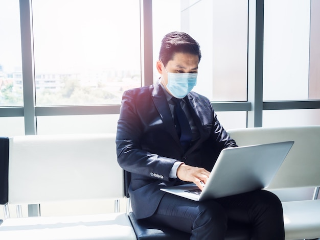 Asian businessman in suit wearing protective face mask using laptop computer on his lap while sitting in modern office building near huge glass window.