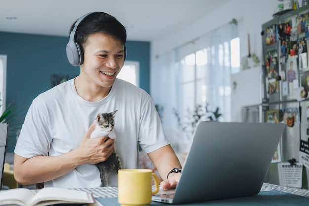 Asian businessman talking to colleague team in video call conference with kitten and smile face. Man using computer laptop and headphone for online meeting. Smart working from home concept.