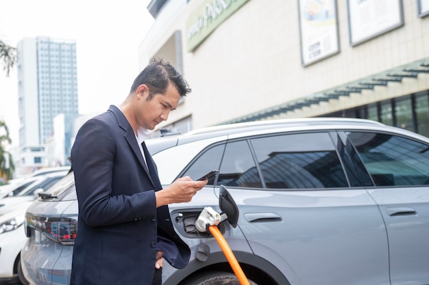 Asian businessman traveling with electric car stopping at charging station standing plugged in internet cable on smartphone smiling joyfully while charging energy saving electric car view