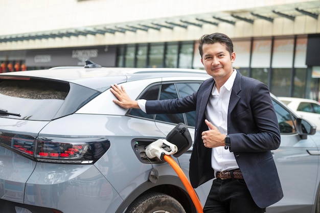 アジア系のビジネスマンが充電ステーションに立って電気自動車を持って旅行し、スマートフォンのインターネットケーブルに差し込まれ、エネルギーを節約する電気自動車のビューを充電しながら楽しそうに微笑む