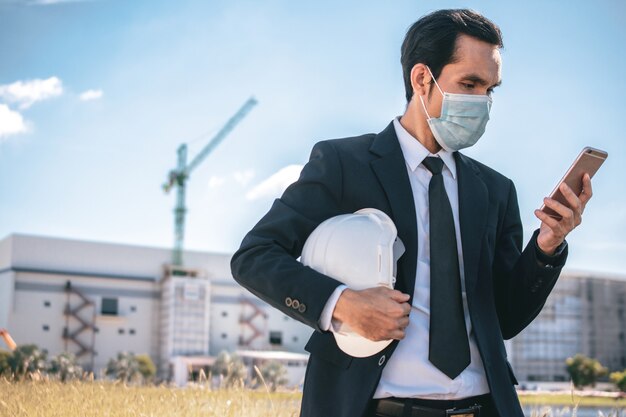 L'uomo d'affari asiatico indossa la maschera per il viso chiama il telefono all'aperto in cantiere