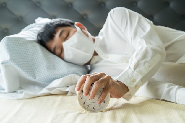 Asian businessman wearing surgical mask sleeping on the bed with alarm clock