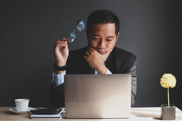 Азиатский бизнесмен в костюме держит очки и просматривает бизнес-отчеты на ноутбуке с кофе