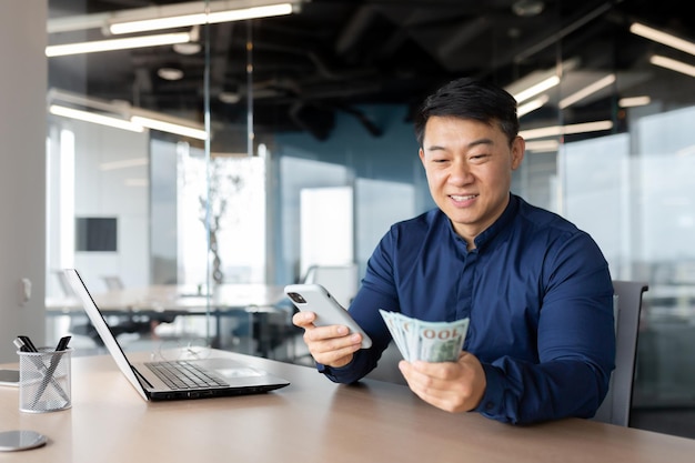 Фото Азиатский бизнесмен выиграл долларовые деньги, мужчина сидит в офисе с наличными и мобильным телефоном