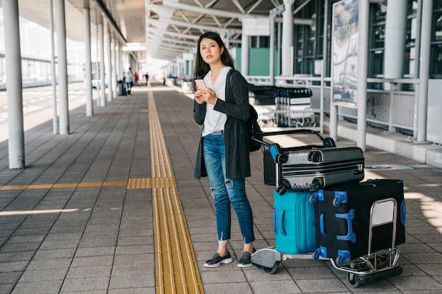 사진 아시아 여성 사업가가 간사이 국제공항에 도착하여 야외에 서서 휴대폰으로 온라인으로 버스 시간표를 검색합니다. 옆에 수하물로 운송을 기다리는 젊은 여자. 해외 생활.