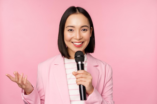 Asian businesswoman giving speech holding microphone and smiling standing in suit over pink background