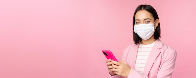 Азиатская деловая женщина в медицинской маске с помощью мобильного телефона японская продавщица корпоративная леди в костюме, держащая смартфон, стоящий на розовом фоне