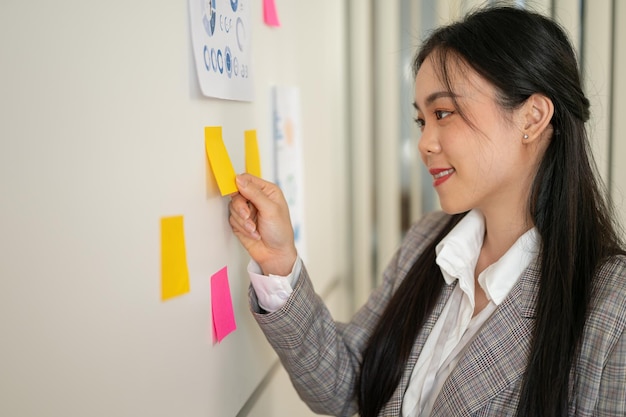 An Asian businesswoman is attaching a sticky note to the wall and sharing her idea with her team