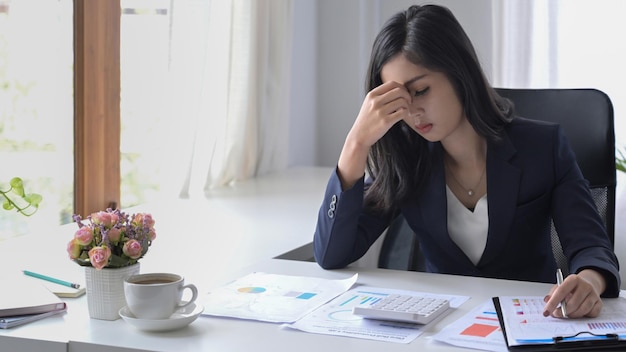 Donna d'affari asiatica che sembra preoccupata stanca e sopraffatta mentre lavora alla scrivania dell'ufficio