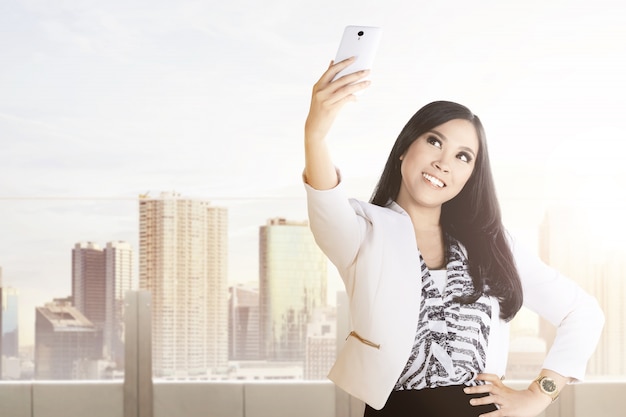 Азиатская коммерсантка делая selfie используя камеру ее телефона на террасе офиса