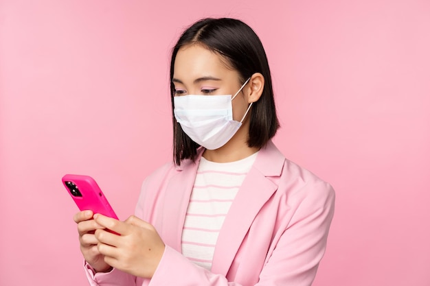 Asian businesswoman in medical face mask using mobile phone Japenese saleswoman corporate lady in suit holding smartphone standing over pink background