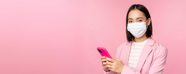 Asian businesswoman in medical face mask using mobile phone Japenese saleswoman corporate lady in suit holding smartphone standing over pink background