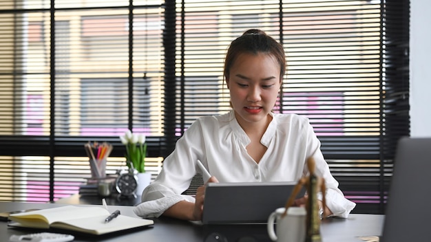 Asian businesswoman sitting at modern workplace and using digital tablet