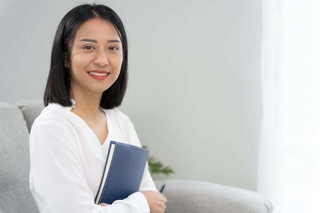 Азиатская деловая женщина улыбается и держит книгу в офисе Красивая и симпатичная азиатка сидит на диване женские портреты