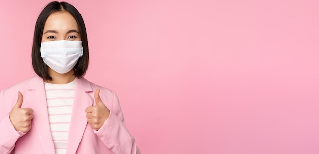 Asian businesswoman in suit and medical face mask showing thumbs up recommending wearing personal protective equipment in office during covid19 pandemic pink background