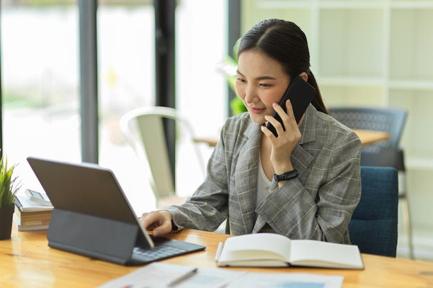 Donna d'affari asiatica che parla al telefono cellulare lavorando su tablet portatile nell'area di lavoro dell'ufficio, negoziando cellulare con cliente aziendale o cliente