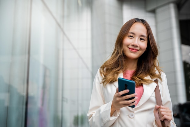 Азиатская деловая женщина пишет смс на смартфоне, добираясь на работу, она идет рядом со своим офисным зданием, счастливая деловая женщина улыбается с помощью мобильного телефона на улице, гуляя по городской улице, смотрит в камеру