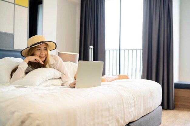 Asian businesswoman traveller in suit in hotel room with travel luggage sitting on bed using computer laptop