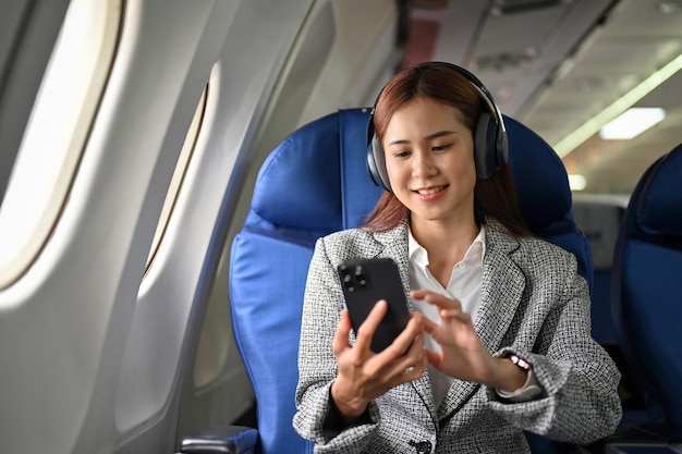 飛行機で旅行中にヘッドフォンで音楽を聴いてスマートフォンを使用してアジアの実業家
