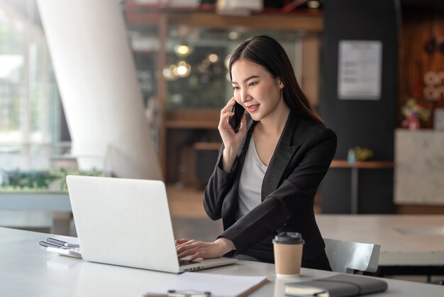 La donna di affari asiatica che lavora ad un colloquio del laptop sul telefono con i clienti all'ufficio.