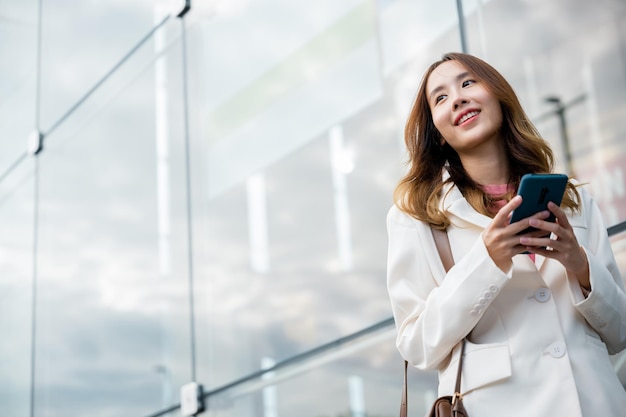 Азиатская деловая женщина, работающая с мобильным телефоном, стоящая напротив уличного здания возле офиса, Портрет успешной деловой женщины, улыбающейся, держащей смартфон, использующий онлайн-чат по утрам