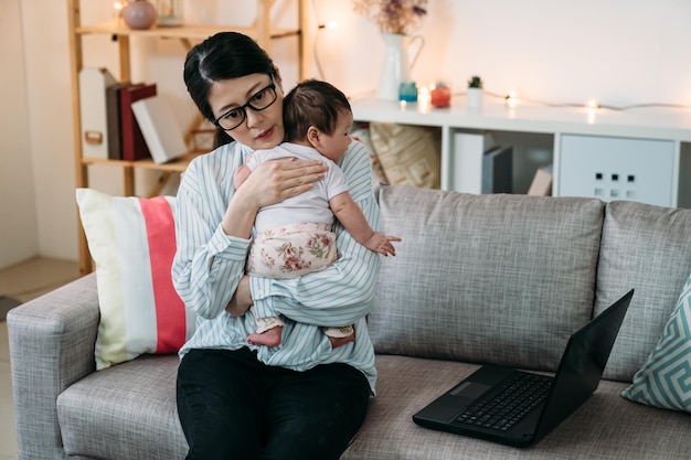 asian career mom is hugging and comforting her baby child while working from home with a laptop on the sofa in the living room.