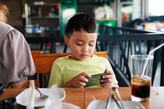 Ragazzo asiatico del bambino che gioca online sullo smartphone al ristorante mentre aspetta il cibo alla griglia della mamma