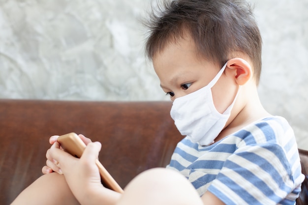 Il ragazzo asiatico del bambino che indossa una maschera di protezione gioca sullo smartphone di casa in quarantena dal coronavirus covid-19 e dall'inquinamento atmosferico pm2.5.